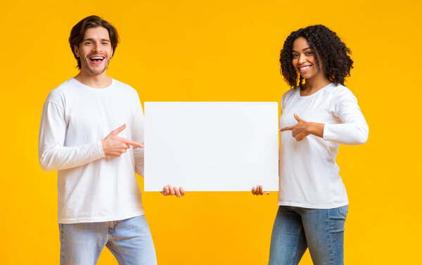 Joyful interracial couple pointing at white blank placard in their hands