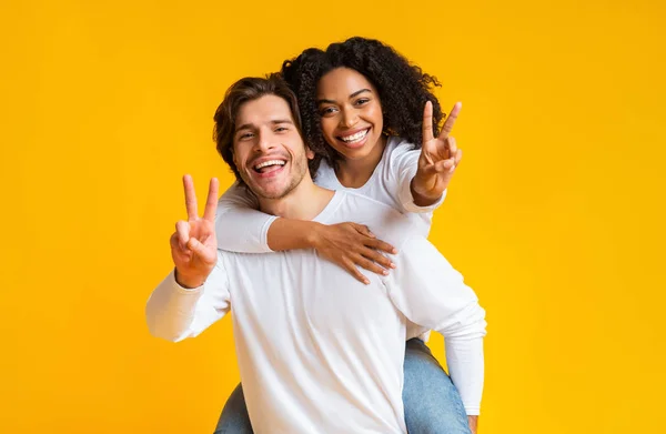 Gioiosa coppia interrazziale abbracciando e mostrando segno di pace su sfondo giallo — Foto Stock