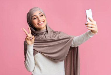 Neşeli Müslüman kadın akıllı telefondan selfie çekiyor ve barış jesti gösteriyor.