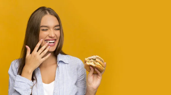 Ragazza felice mangiare hamburger ridere su sfondo giallo Studio, Panorama — Foto Stock