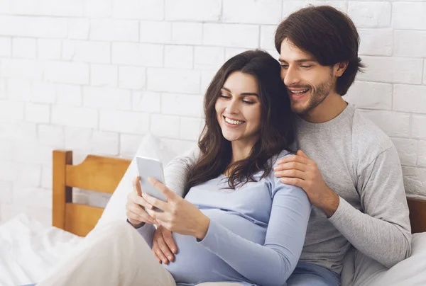 Feliz pareja embarazada posando para selfie, abrazándose en la cama en casa —  Fotos de Stock