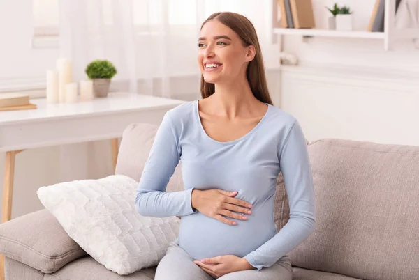 Mãe-To-Be feliz tocando barriga grávida sentada no sofá interior — Fotografia de Stock