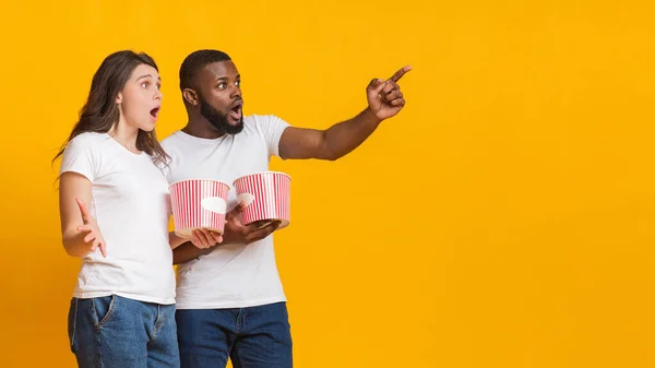 Sorprendidos amigos interracial sosteniendo cubos de palomitas de maíz y apuntando al espacio de copia —  Fotos de Stock