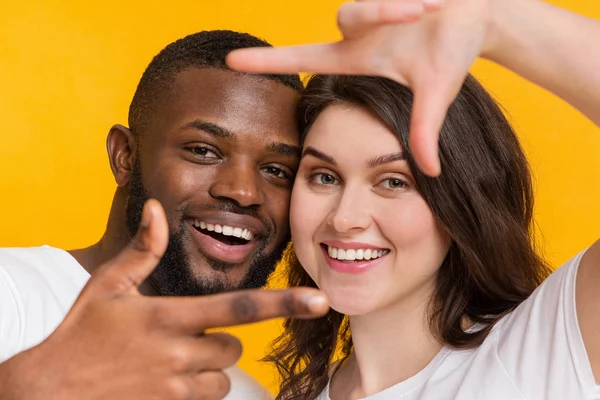 Casal multirracial positivo olhando através da moldura do dedo, posando sobre fundo amarelo — Fotografia de Stock