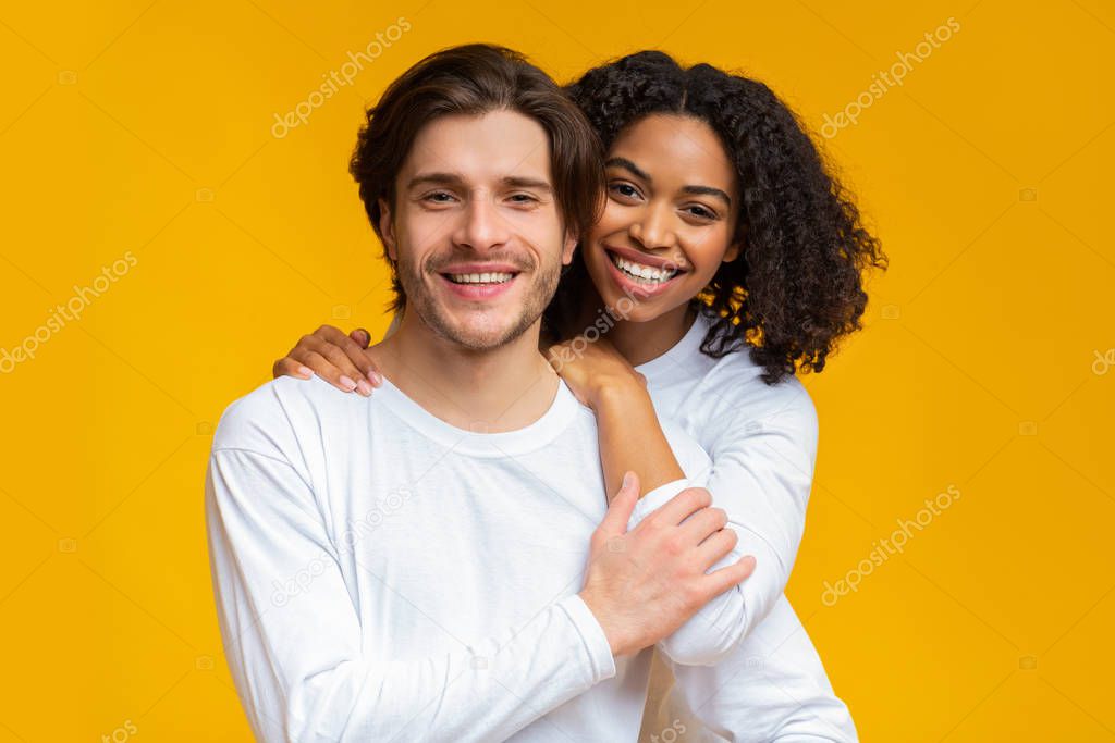 Romantic interracial couple embracing and posing over yellow background