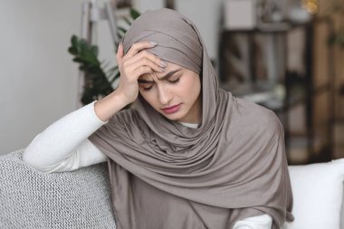 Başörtülü depresif Müslüman kadın koltukta oturuyor ve alnına dokunuyor.