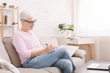 Emekli kadın evde dizüstü bilgisayar kullanarak not yazıyor.