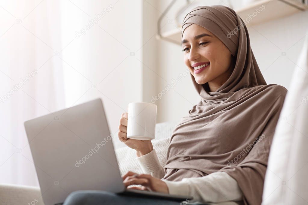 Muslim Woman In Hijab Using Laptop And Drinking Coffee At Home