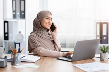 Başörtülü Müslüman İş kadını ofiste çalışırken cep telefonuyla konuşuyor