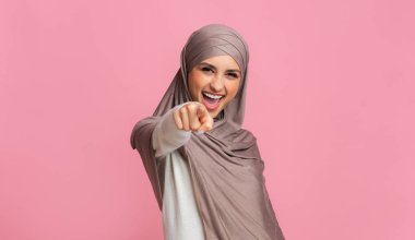 Arap kadın gülüyor ve pembe arka planda kamerayı işaret ediyor.