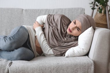 Evde karın ağrısı çeken tesettürlü mutsuz bir Arap kadın.