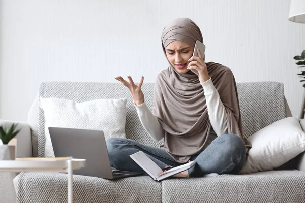 Muchacha musulmana preocupada hablando en el teléfono celular mientras usa el ordenador portátil en casa — Foto de Stock
