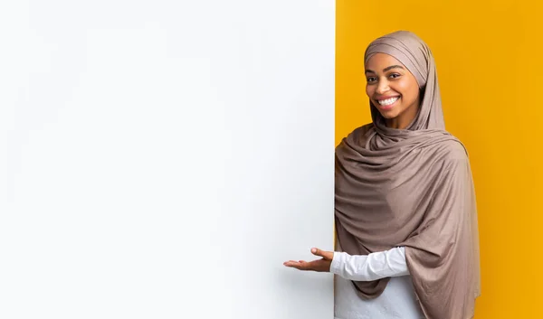 Usmívající se afro muslimka v šátku ukazující na bílou reklamní tabuli — Stock fotografie