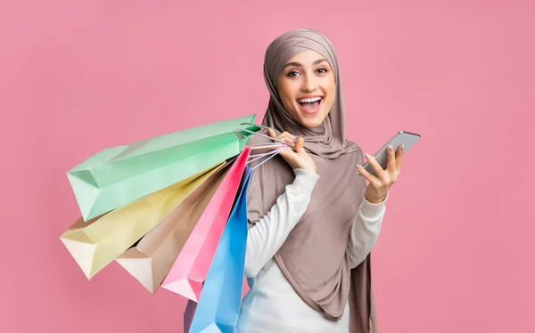 Wanita arab ceria berkerudung memegang smartphone dan kantong kertas cerah — Stok Foto