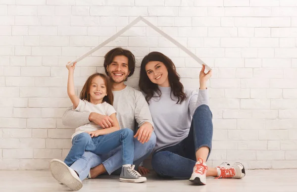 Szczęśliwa rodzina siedzi pod symbolicznym dachem marząc o nowym domu. — Zdjęcie stockowe