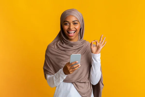 Chica musulmana negra en hijab usando un teléfono inteligente y mostrando un gesto aceptable —  Fotos de Stock