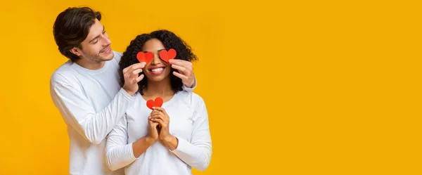 Romantique gars couvrant les yeux de sa petite amie avec des coeurs de papier rouge — Photo