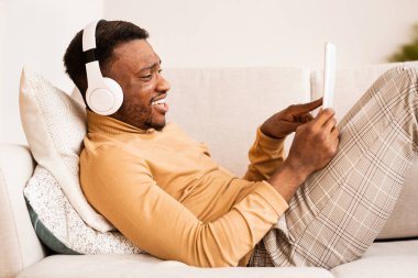 Kulaklık takan mutlu adam tablet kullanıyor. Kapalı koltukta yatıyor.