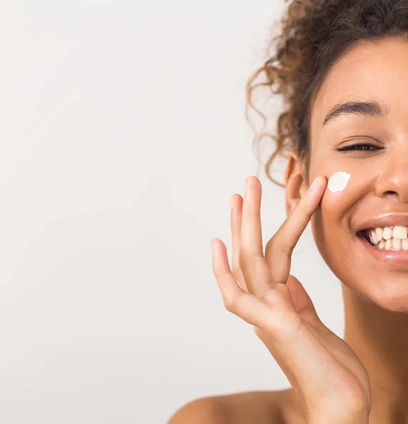 Cuidado com o rosto. Mulher negra feliz aplicando creme hidratante na bochecha — Fotografia de Stock