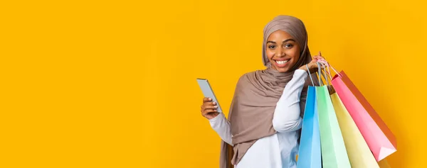 Feliz afro musulmana chica en pañuelo para la cabeza celebración de teléfonos inteligentes y bolsas de compras —  Fotos de Stock