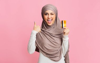 Tesettürlü Müslüman bir kadın kredi kartı tutuyor ve başparmağını kaldırıyor.