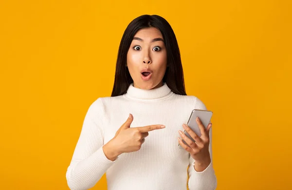 Geschokt aziatisch meisje wijzend naar mobiele telefoon en kijken naar camera — Stockfoto