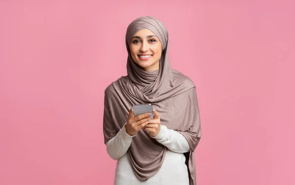 Sonriente chica islámica en pañuelo para la cabeza sosteniendo el teléfono inteligente y mirando a la cámara —  Fotos de Stock