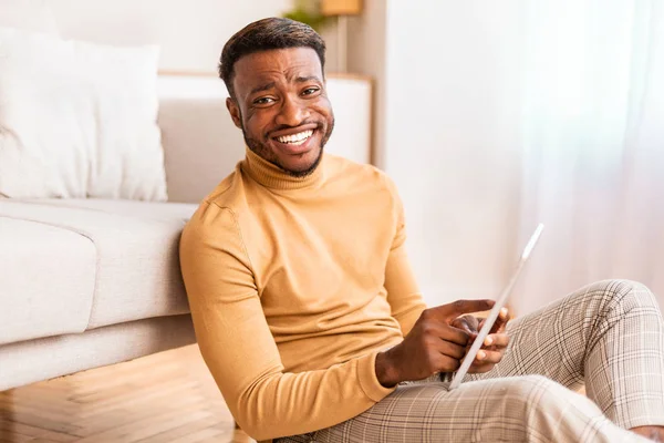Afro άνθρωπος χρησιμοποιώντας Tablet χαμογελώντας κάθεται στο πάτωμα στο σπίτι — Φωτογραφία Αρχείου