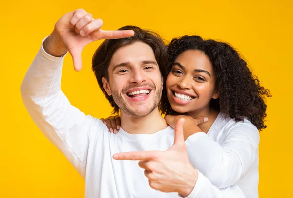 Casal multirracial positivo olhando através da moldura do dedo, posando sobre fundo amarelo — Fotografia de Stock