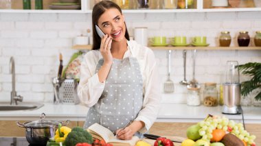 Woman taking down recipe, talking on phone in kitchen clipart