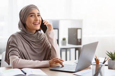 Mutlu İslamcı İş kadını cep telefonuyla konuşuyor ve ofiste dizüstü bilgisayar kullanıyor.