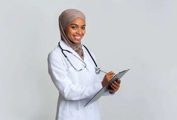 Lächelnde schwarze muslimische Ärztin im Hijab schreibt Notizen an Klemmbrett — Stockfoto