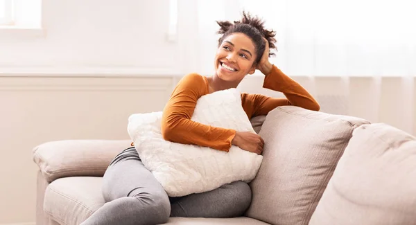 Afroamerikanerin sitzt auf Sofa und umarmt Kissen drinnen, panorama — Stockfoto