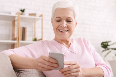 Neşeli yaşlı bayan evde modern cep telefonunun keyfini çıkarıyor.