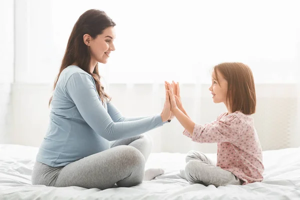 Adorable niña jugando con su madre embarazada, sentada en la cama —  Fotos de Stock