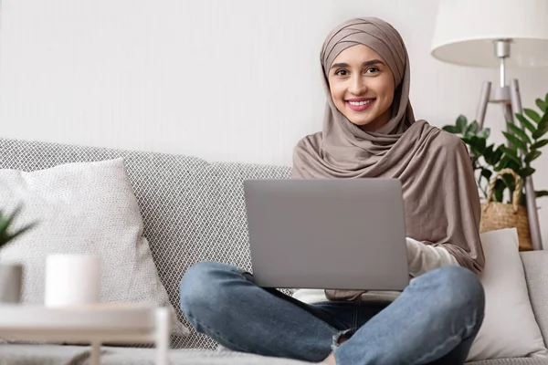 Muslim gadis dalam jilbab duduk di sofa dengan laptop di lap — Stok Foto