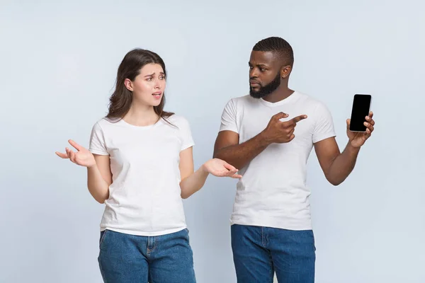 Ревнивый черный парень держит смартфон, спрашивает объяснения у своей изменяющей подружки — стоковое фото