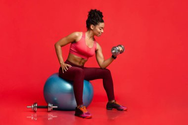 Fitness modeli ağırlık pompalıyor, fitness topunun üzerinde oturuyor.