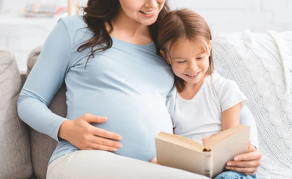 Chica feliz divirtiéndose mientras lee libro con mamá embarazada — Foto de Stock