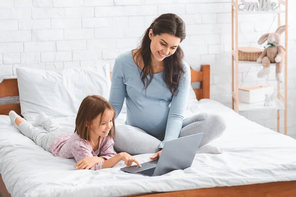Mała córka i ciężarna matka za pomocą laptopa, siedzi na łóżku — Zdjęcie stockowe