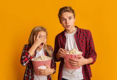 Scared brother and sister holding popcorn buckets and looking at camera clipart