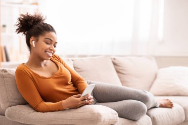 Kanepede oturan akıllı telefondan müzik dinleyen Afro Kadın.