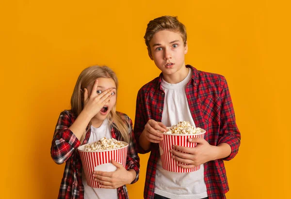 Scared brother and sister holding popcorn buckets and looking at camera — Stock Photo, Image