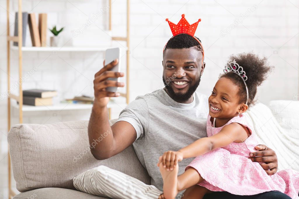 Happy african father and daughter wearing crowns, taking selfie