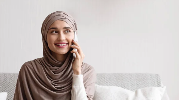Fiatal muszlim nő fejkendőben, kellemes telefonbeszélgetést folytat otthon — Stock Fotó