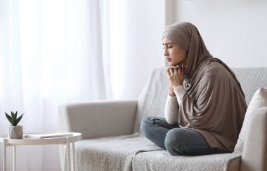 Başörtülü depresif Müslüman kadın evdeki kanepede üzgün oturuyor.