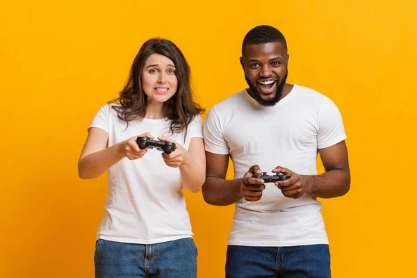 Casal multirracial de jogos de azar jogando videogames com joysticks, posando sobre fundo amarelo — Fotografia de Stock