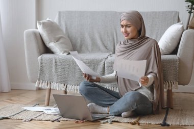 Kağıtlar ve dizüstü bilgisayarlarla endişe içinde oturan mutsuz bir Arap kadın.