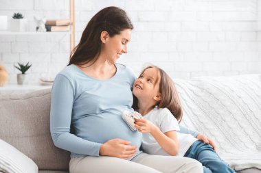 Küçük kız annesinin karnını beklerken bebek botları giyiyor.