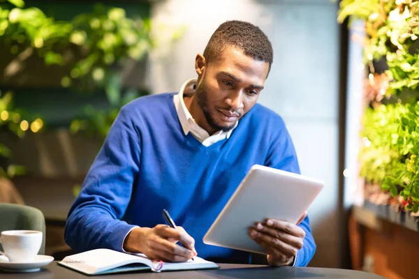 Hombre negro sosteniendo la tableta y escribiendo en un cuaderno —  Fotos de Stock